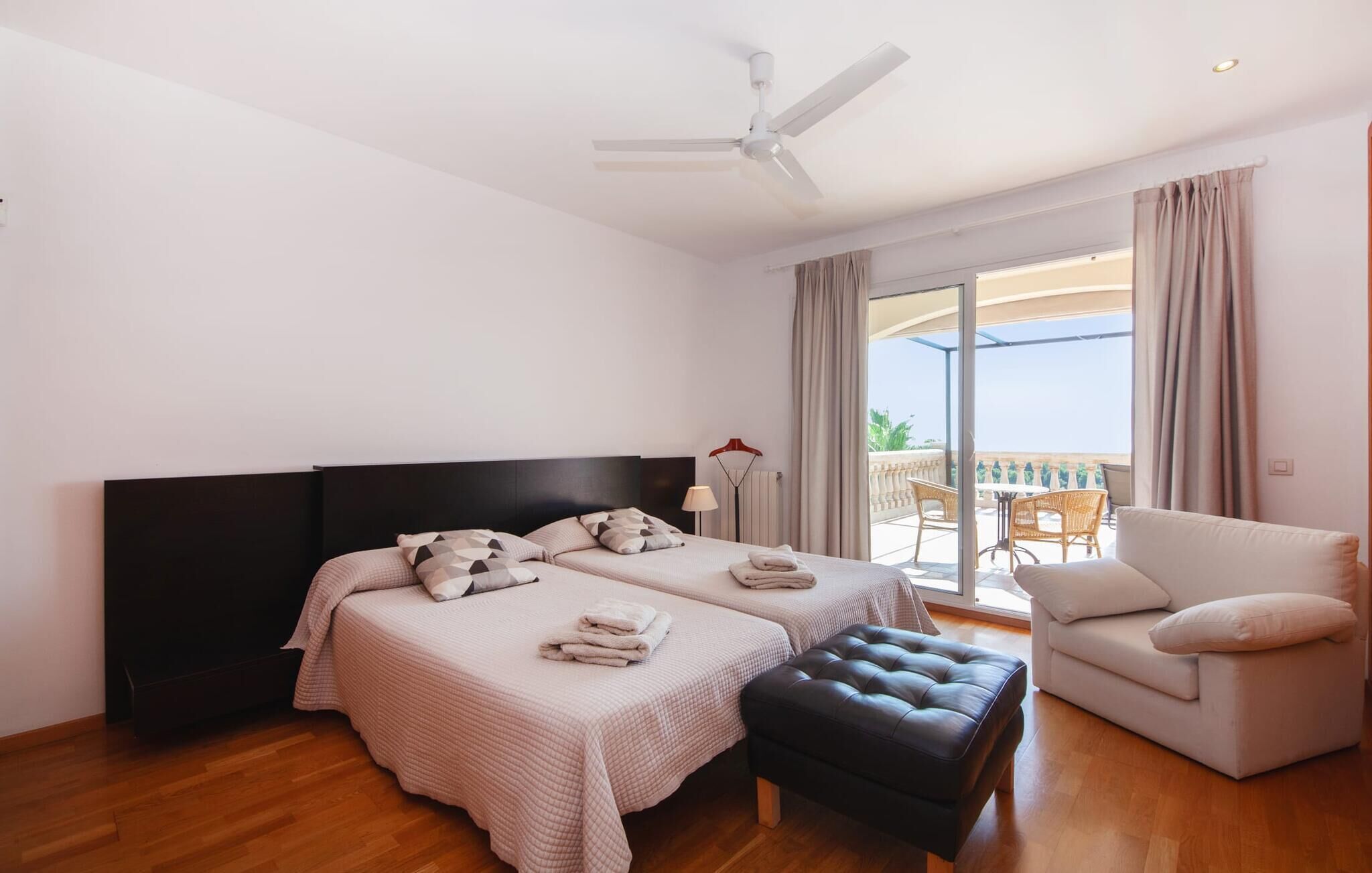 A hotel room with two beds, a ceiling fan, a balcony with outdoor seating, and a view of the sea.