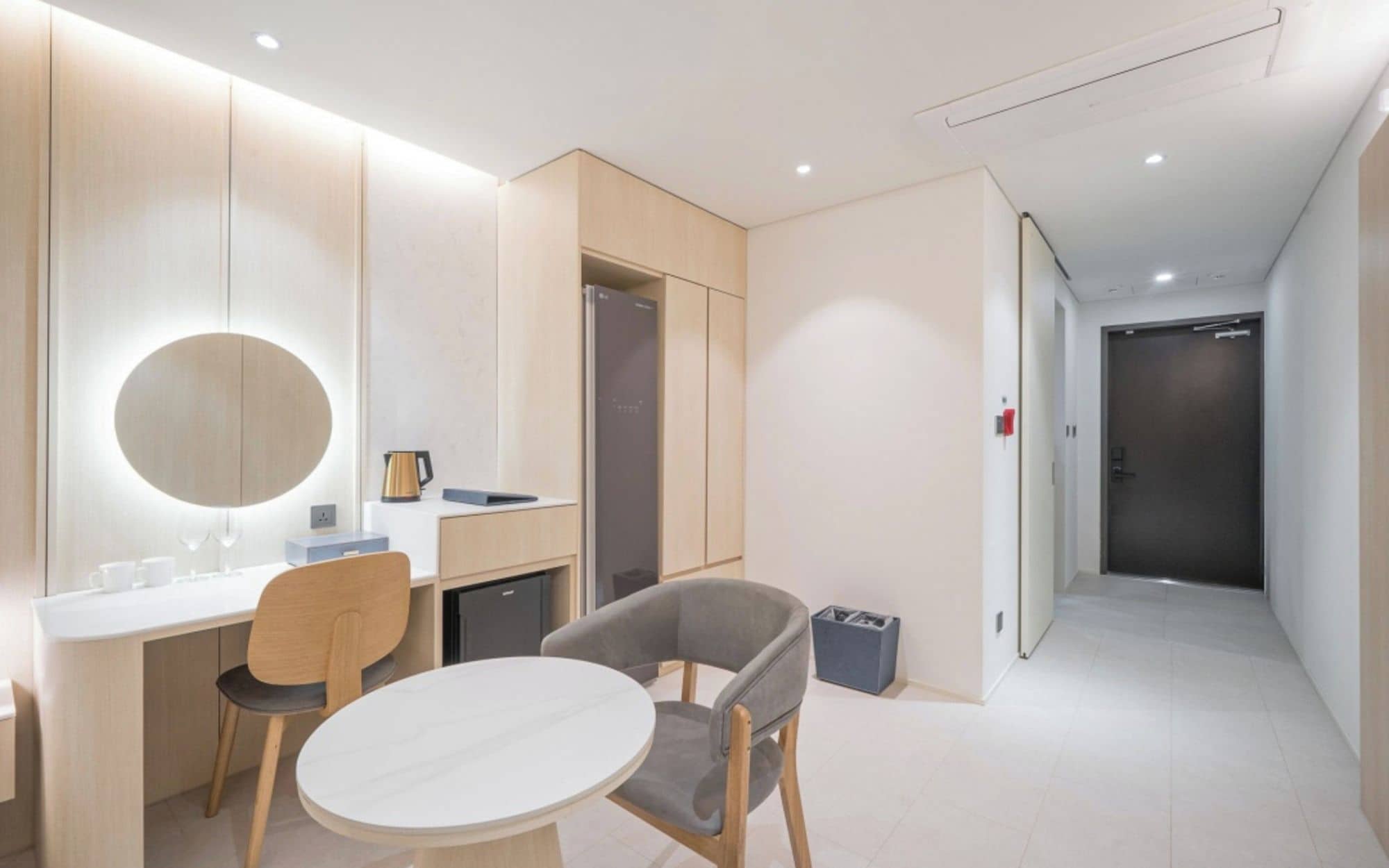 A modern hotel room with a round mirror, a small table, two chairs, a kitchenette, and a door leading to another room.