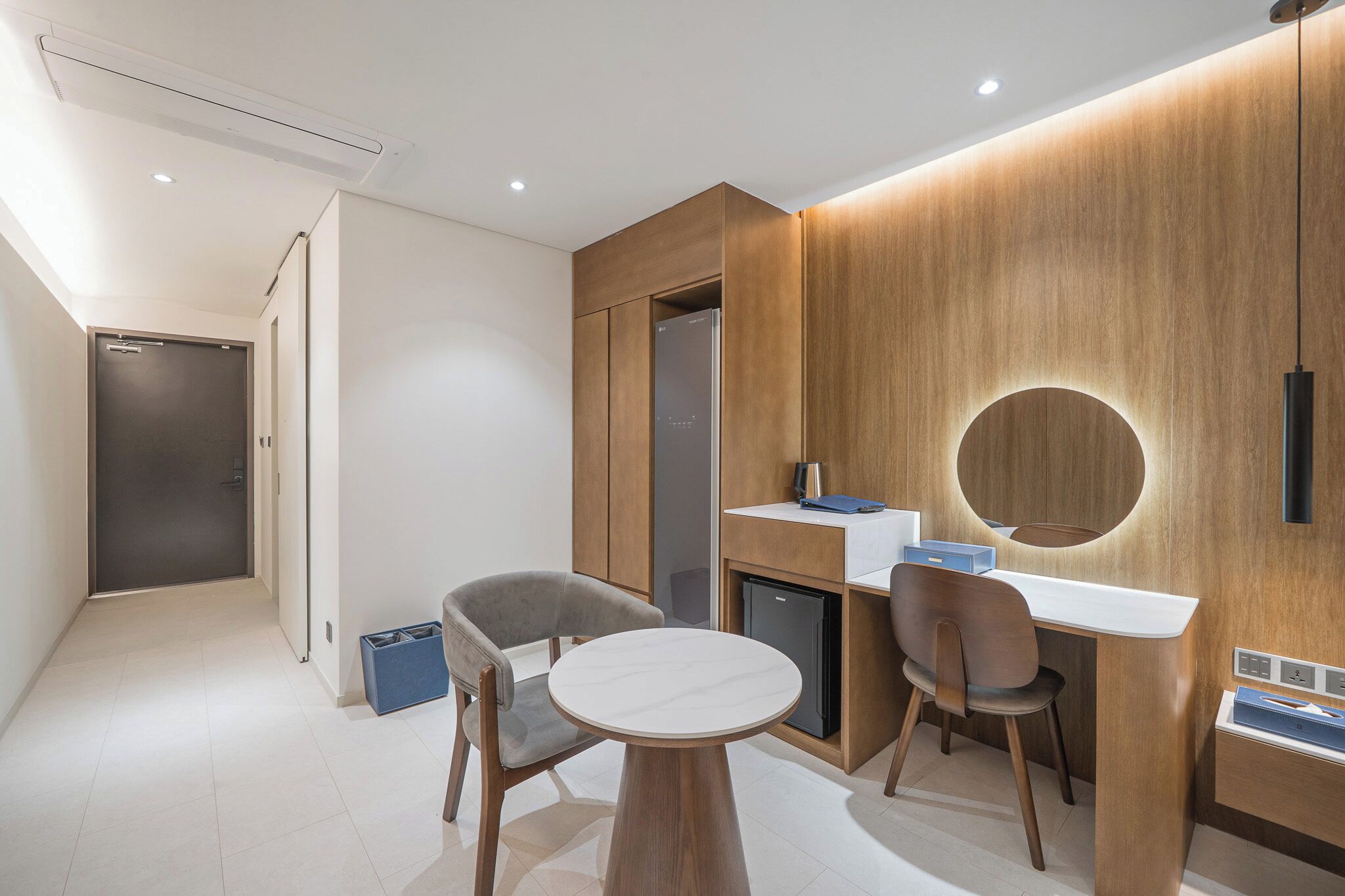 A modern hotel room with a wooden accent wall, a round mirror, a small desk, two chairs, and a compact refrigerator.
