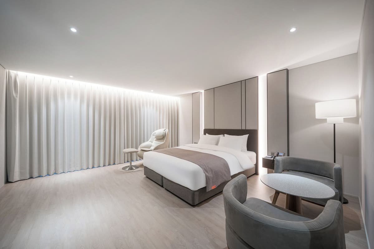 A modern hotel room with a bed, a chair, a small table, and a lamp.