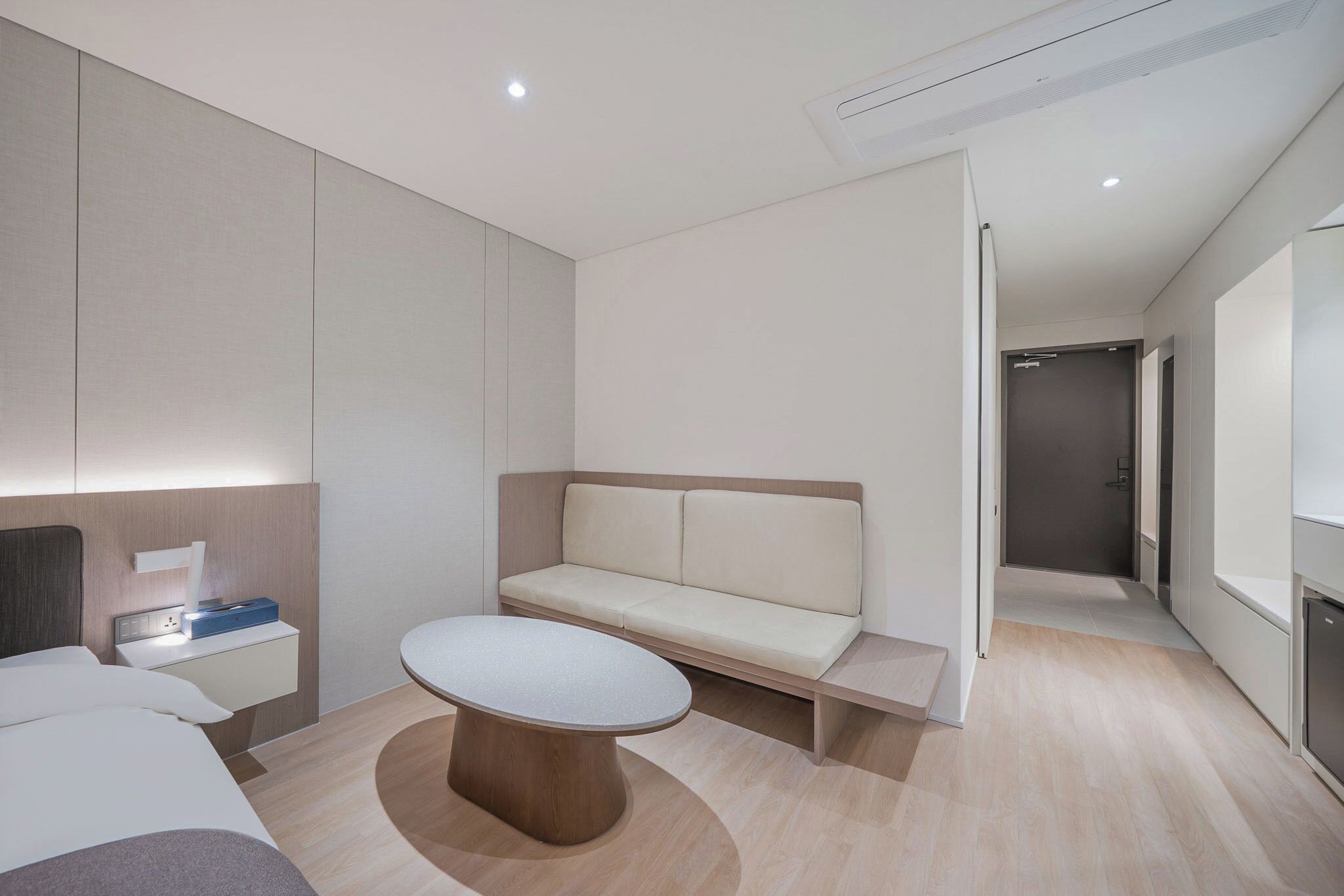 A modern hotel room with a bed, bedside table, a round table, a sofa, and a door leading to another room.