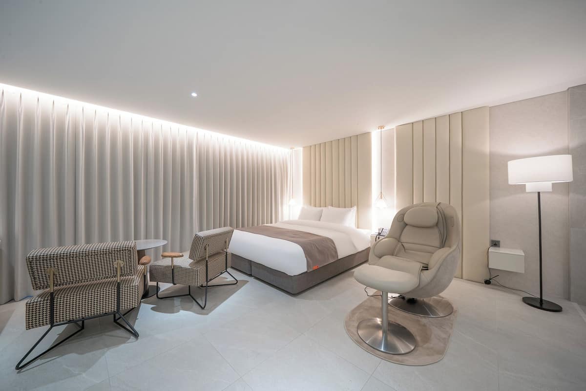 A modern hotel room with a bed, two armchairs, a small table, and a floor lamp.