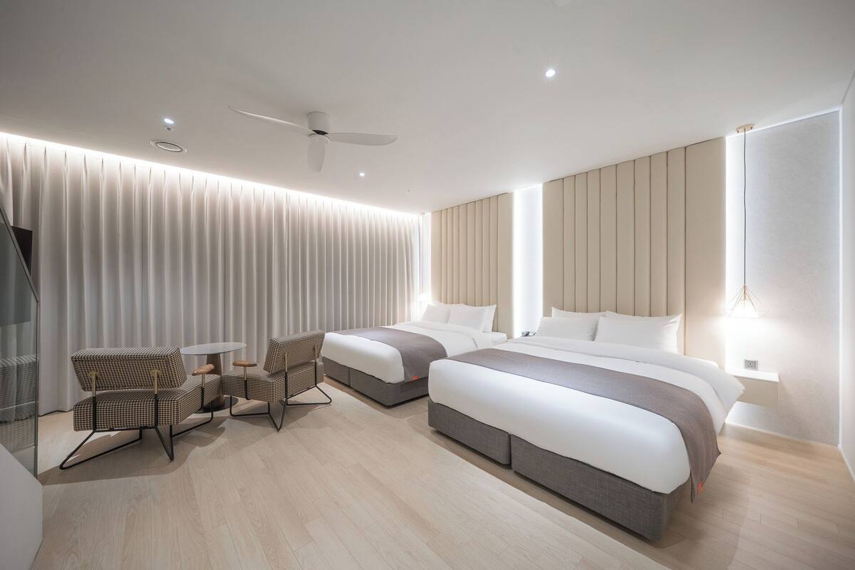 A modern hotel room with two beds, a seating area, and a ceiling fan.