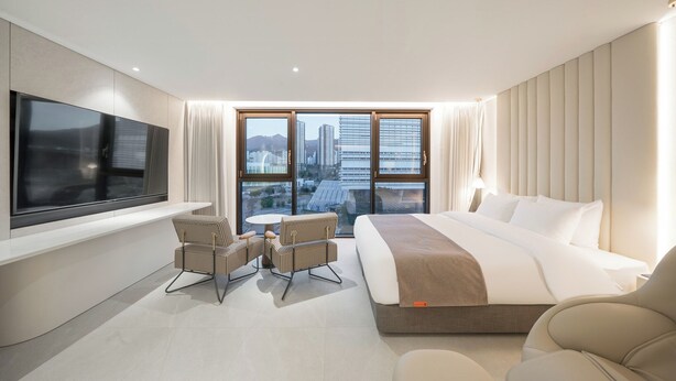 Habitación de hotel moderna con una cama grande, televisión, zona de estar con sillas y vistas a la ciudad a través de amplios ventanales.