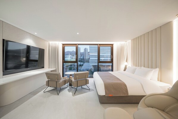 Een moderne hotelkamer met een groot bed, een televisie, een zithoek met stoelen en een stadszicht door grote ramen.