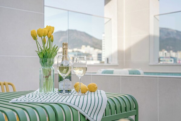 Une table avec une bouteille de champagne, deux verres et des citrons, le tout posé sur une nappe à rayures, avec vue sur une ville et des montagnes.