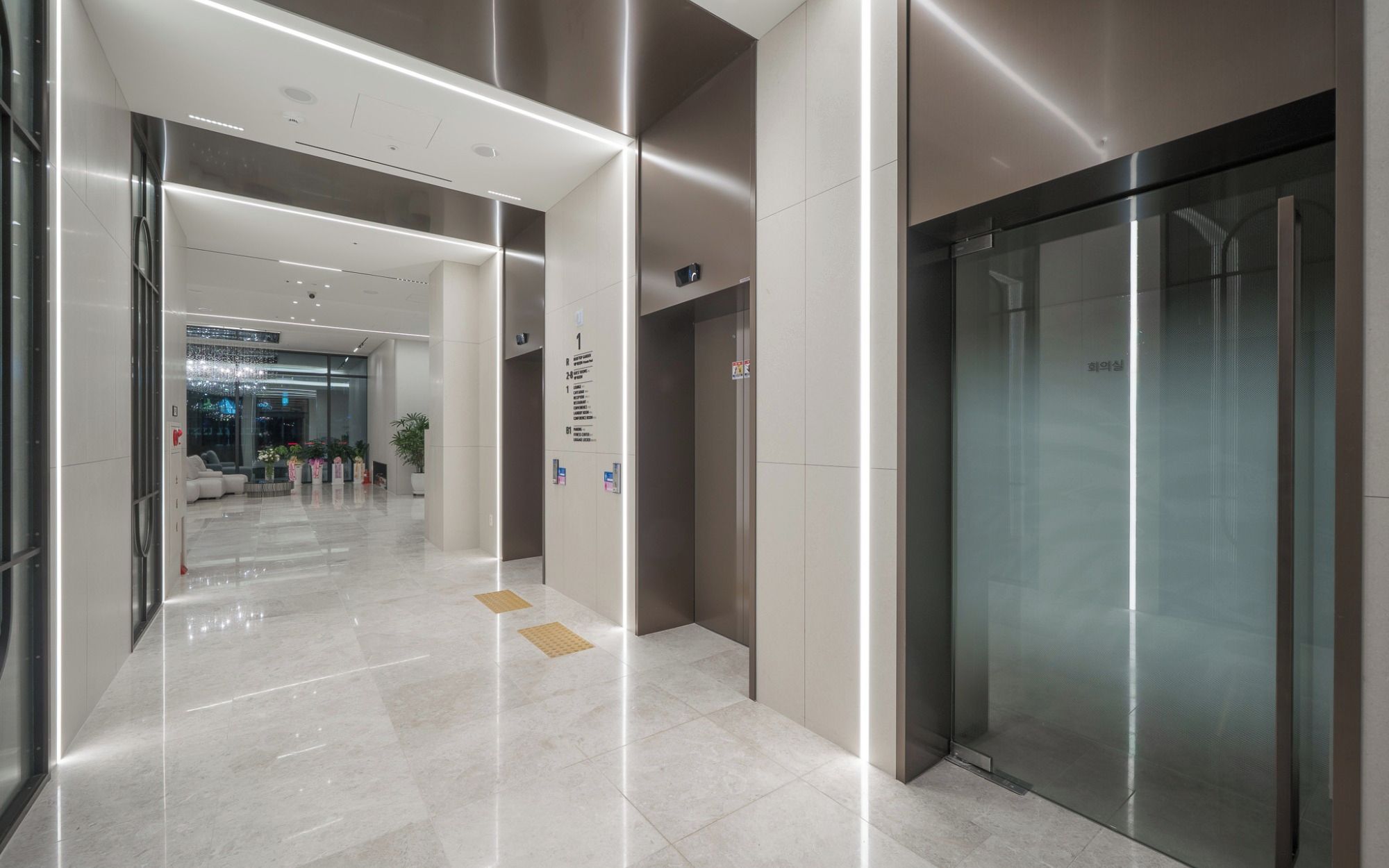 A modern hotel corridor with a glass elevator, a reception area, and a well-lit interior.