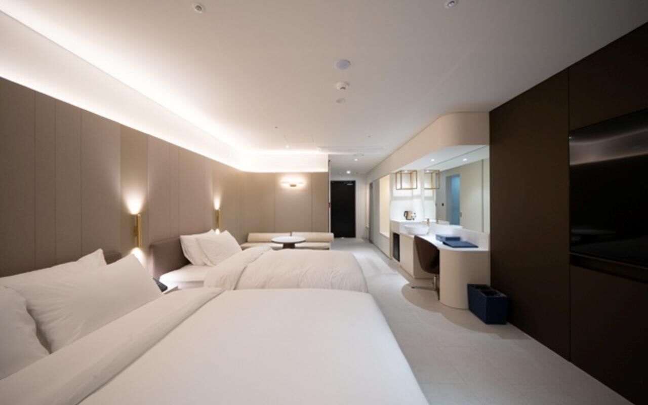 A modern hotel room with two beds, a sitting area, a vanity with a mirror, and a television.