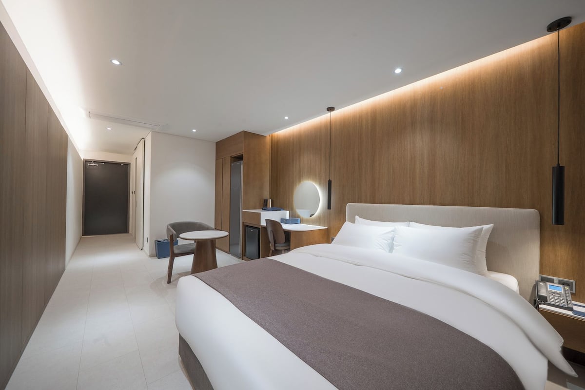 A modern hotel room with a large bed, a desk with a chair, a small round table, a bedside phone, and a mirror.