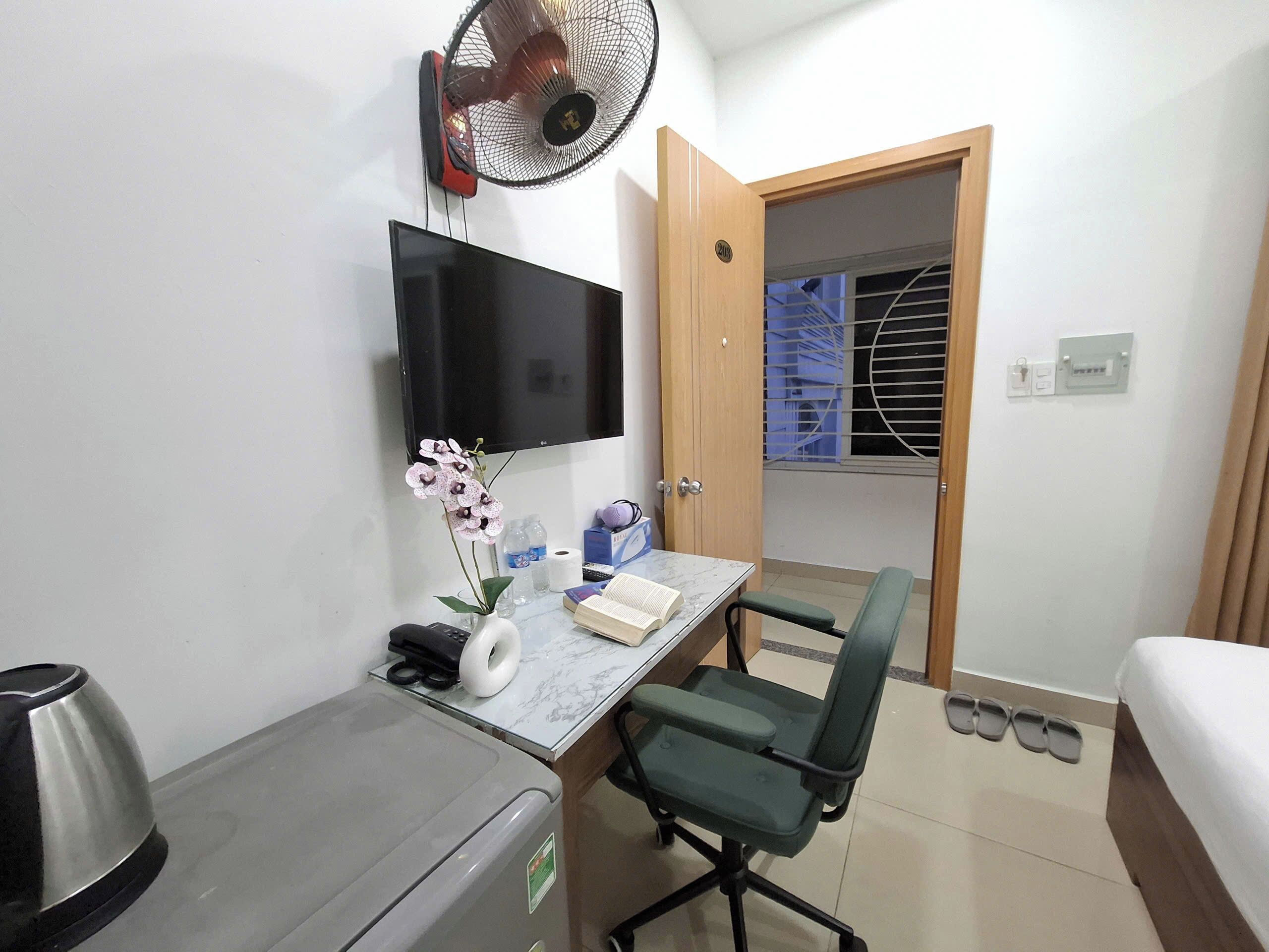 A hotel room with a bed, a desk with a chair, a TV, a fan, a trash bin, and a window with blinds.