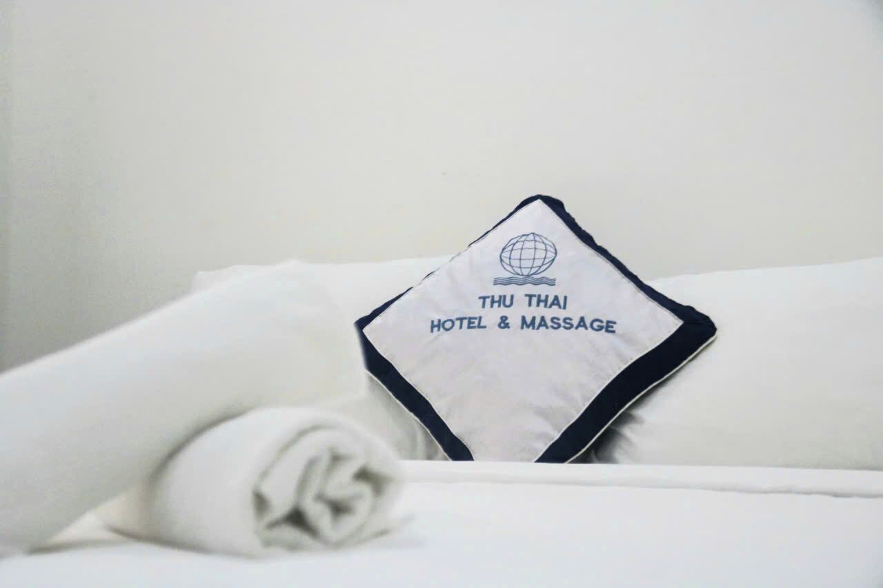 A neatly made bed with a pillow featuring 'THU THAI HOTEL & MASSAGE' text and a globe logo.