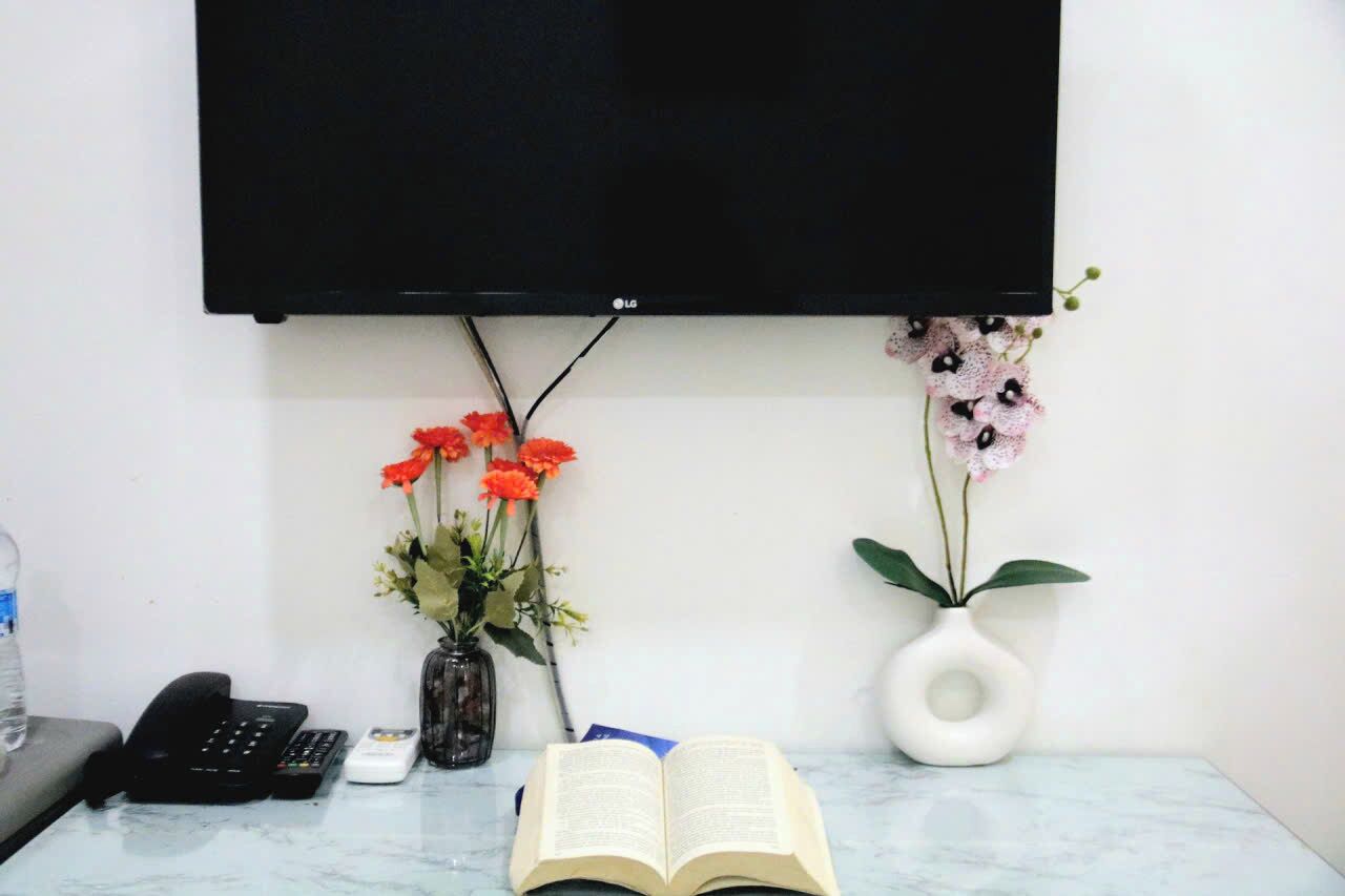 A room with a flat-screen TV mounted on the wall, a telephone, a remote control, an open book, and two vases with flowers.