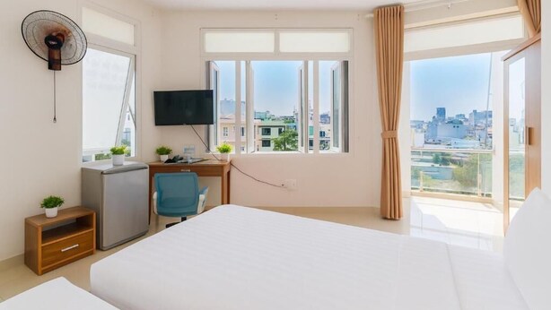 Habitación de hotel con cama, escritorio, silla, televisor y vista a la ciudad a través de ventanas amplias.