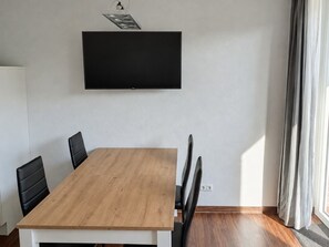 A room with a wooden dining table, black chairs, a flat-screen TV mounted on the wall, and a window with curtains. - "Mour" Vacation Apartment - "Mour" townhouse with a terrace, 2 stories, sleeps 4 (Dahme)