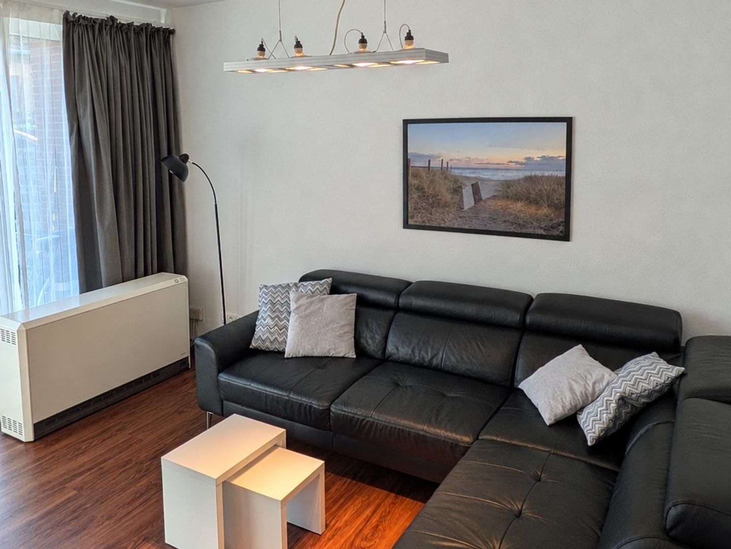 A modern living room with a black leather sofa, a white side table, a floor lamp, and a framed picture on the wall.