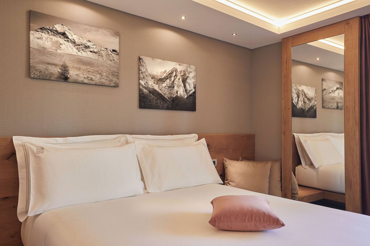 A hotel room with a bed, pillows, and two mountain landscape paintings on the wall.