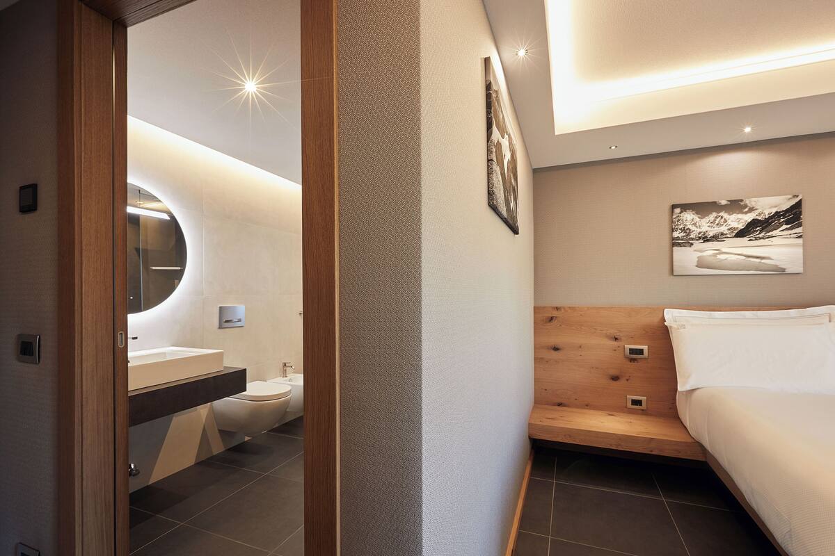 A modern bathroom with a round mirror, a toilet, and a sink. There is a bed with a wooden headboard and a white pillow.