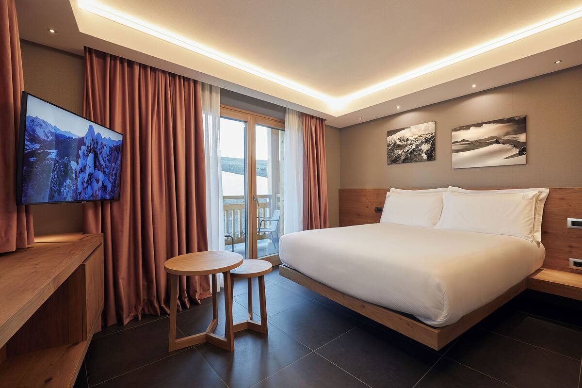 A modern hotel room with a large bed, a TV, a small table, and a balcony with curtains.