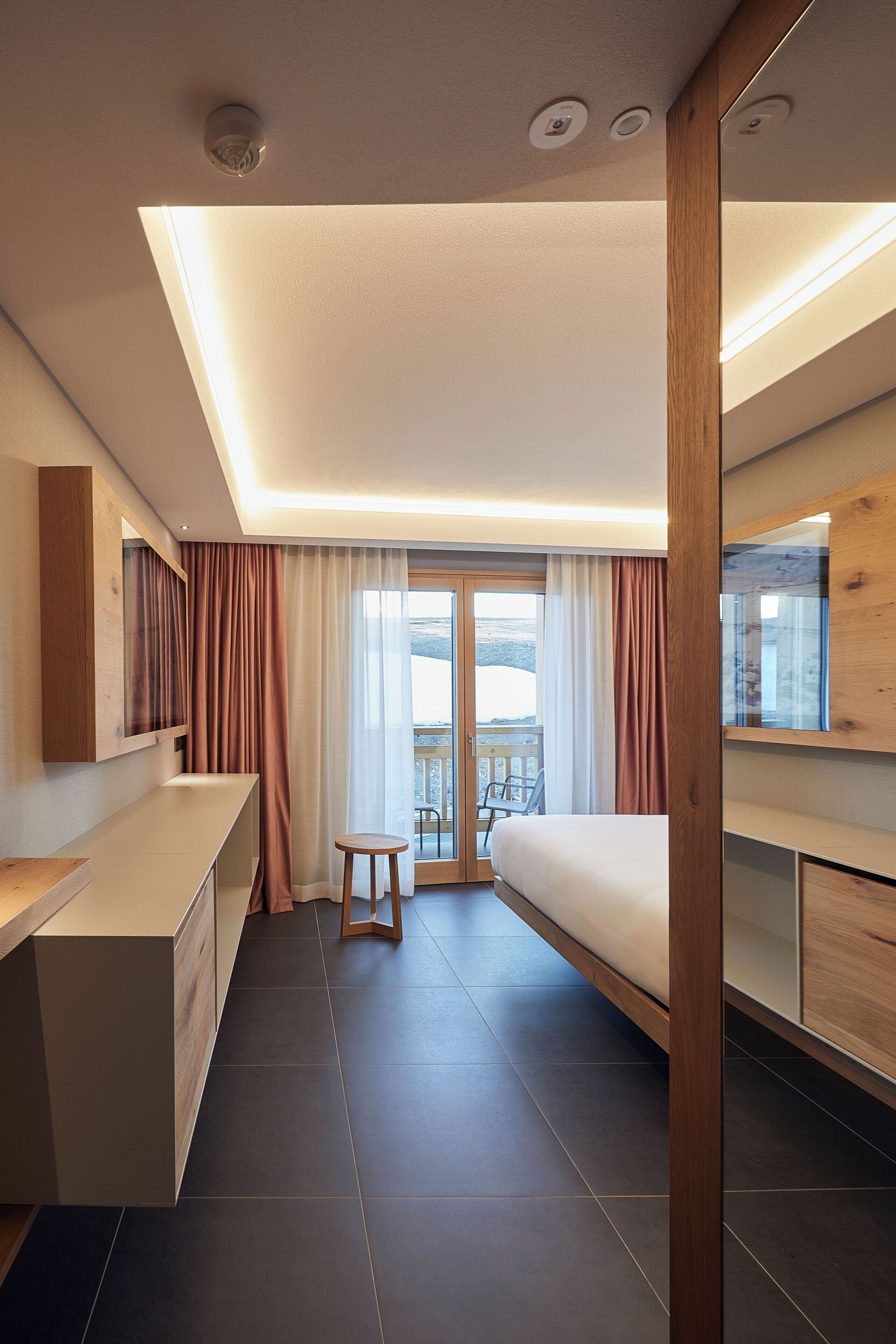 Une chambre d’hôtel moderne, équipée d’un lit, d’un bureau et d’un petit tabouret. Elle dispose également d’un balcon avec vue sur l’extérieur.