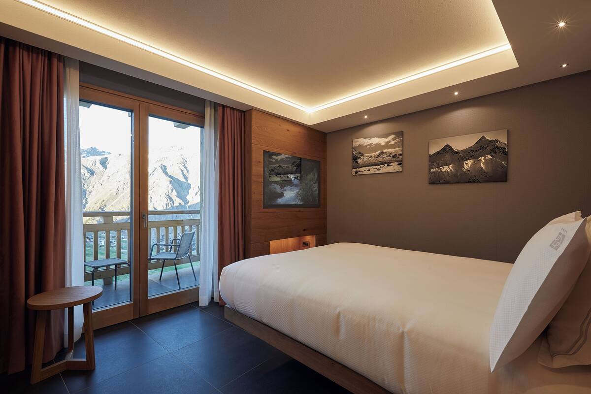 A hotel room with a large bed, a small table, a balcony with a chair, and mountain views.