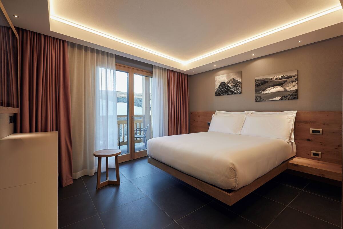 A modern hotel room with a large bed, a small stool, and a view of a mountainous landscape through the window.