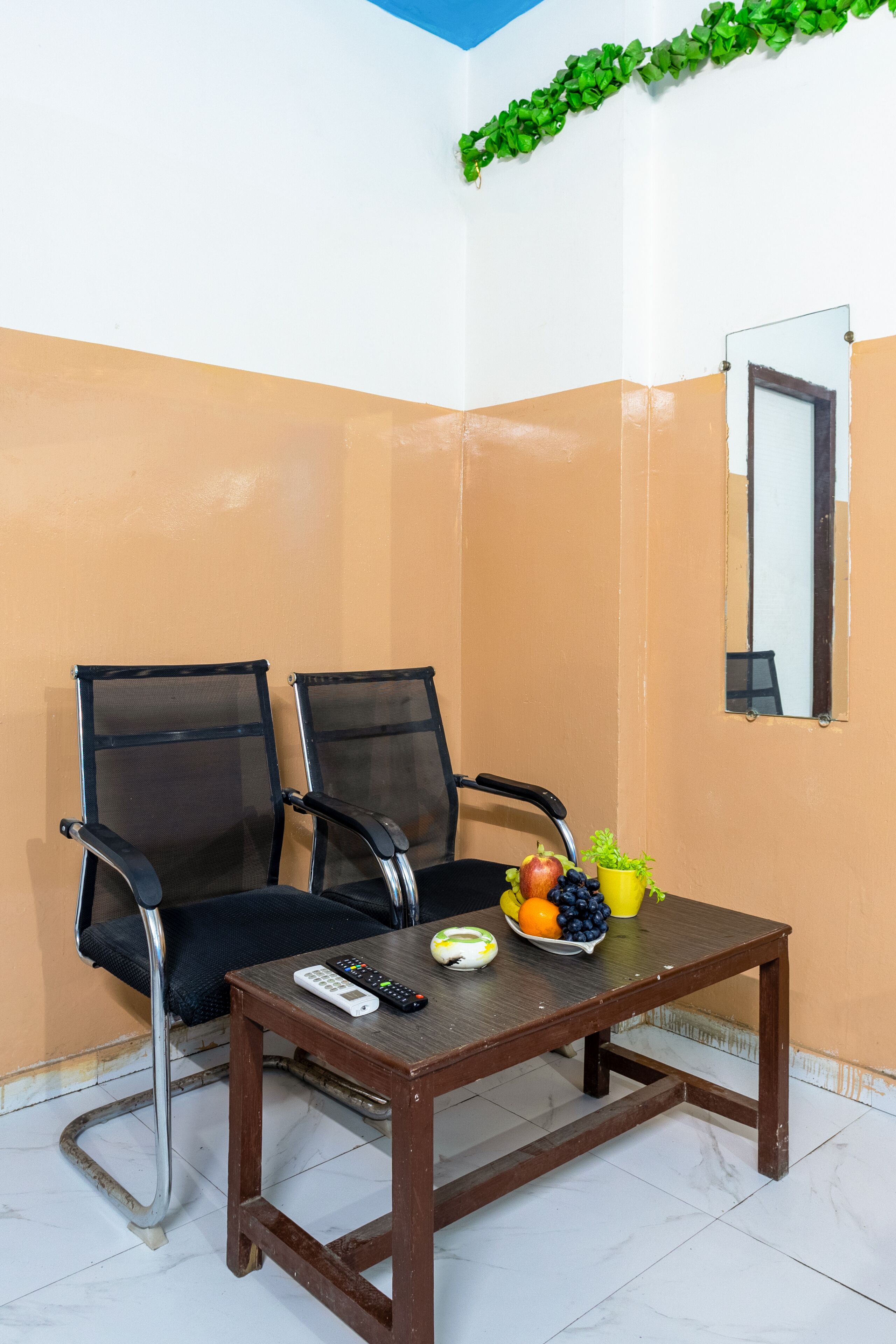 A corner room with two black mesh chairs, a wooden table with a fruit bowl, a remote control, and a mirror on the wall.