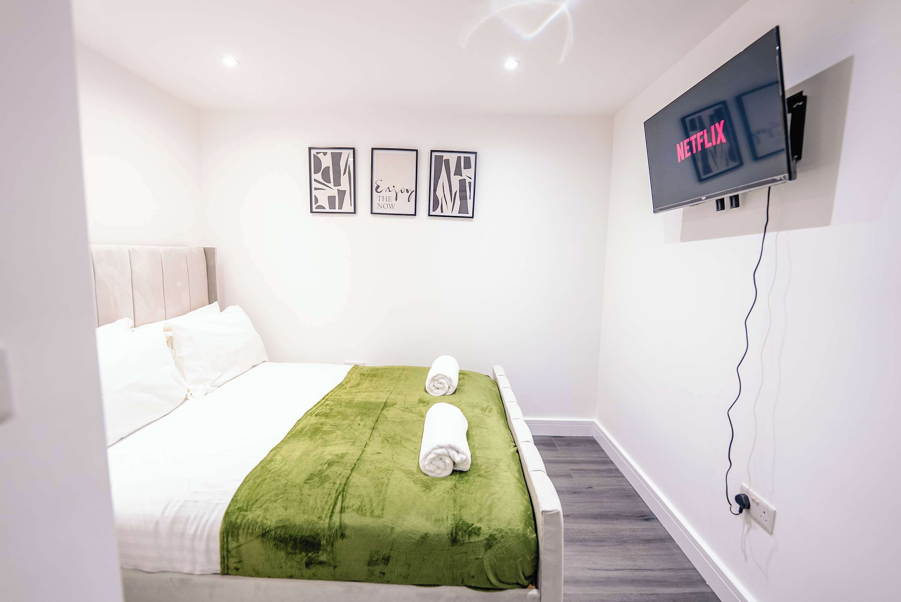 Une chambre d’hôtel avec un lit, une couverture verte, deux serviettes enroulées, un téléviseur fixé au mur qui affiche « NETFLIX », et trois tableaux encadrés accrochés au mur.