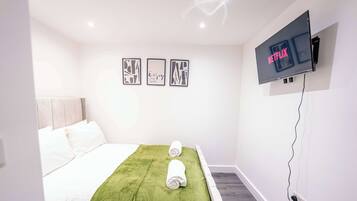 Une chambre d’hôtel avec un lit, une couverture verte, deux serviettes enroulées, un téléviseur fixé au mur qui affiche « NETFLIX », et trois tableaux encadrés accrochés au mur.