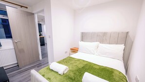 Hotellihuone, jossa on sänky, vihreän ja valkoisen väriset vuodevaatteet, puinen yöpöytä ja valkoinen tuoli.