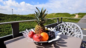 Ein Tisch mit Obstschale, gefüllt mit Äpfeln, einer Ananas, einer Orange und Trauben, auf einem Balkon mit herrlicher Aussicht auf grüne Hügel und einen klaren Himmel.