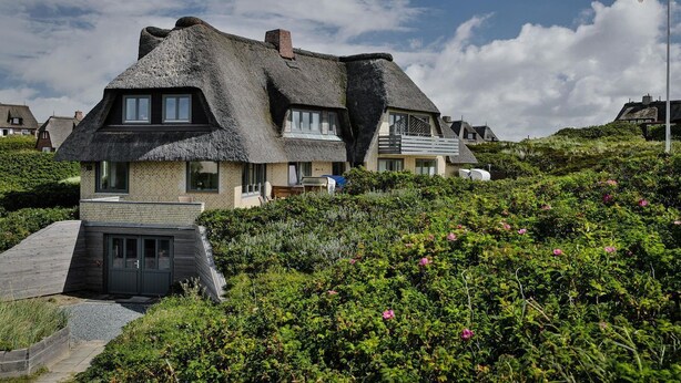 Ein traditionelles Haus mit Strohdach und einem modernen Anbau, umgeben von üppiger Vegetation und einer Flagge auf einem Fahnenmast.