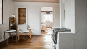 A room with a wooden floor, a white chair, a small table, a bookshelf, a bed with a white bedspread, and a stove. - Cozy Country Home in Quiet Töreboda (Töreboda)