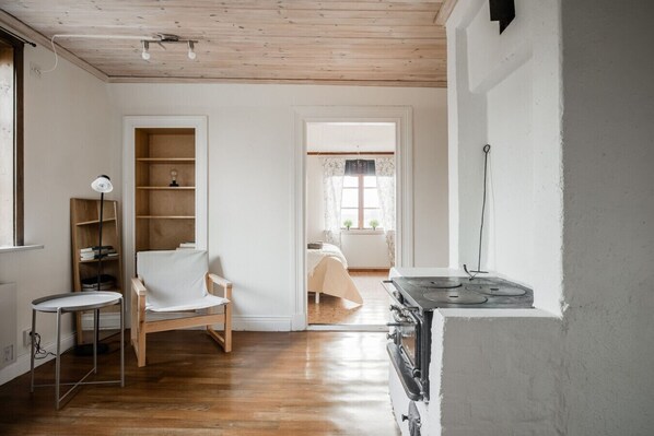 Een kamer met een houten vloer, een witte stoel, een klein tafeltje, een boekenkast, een bed met een witte dekbedovertrek, en een fornuis.