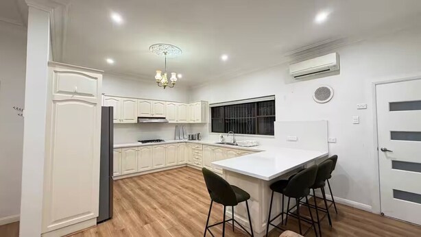 A modern kitchen with white cabinetry, a central island with bar stools, and a built-in oven. - Cozy family-friendly homestay in Bankstown! (Bankstown)