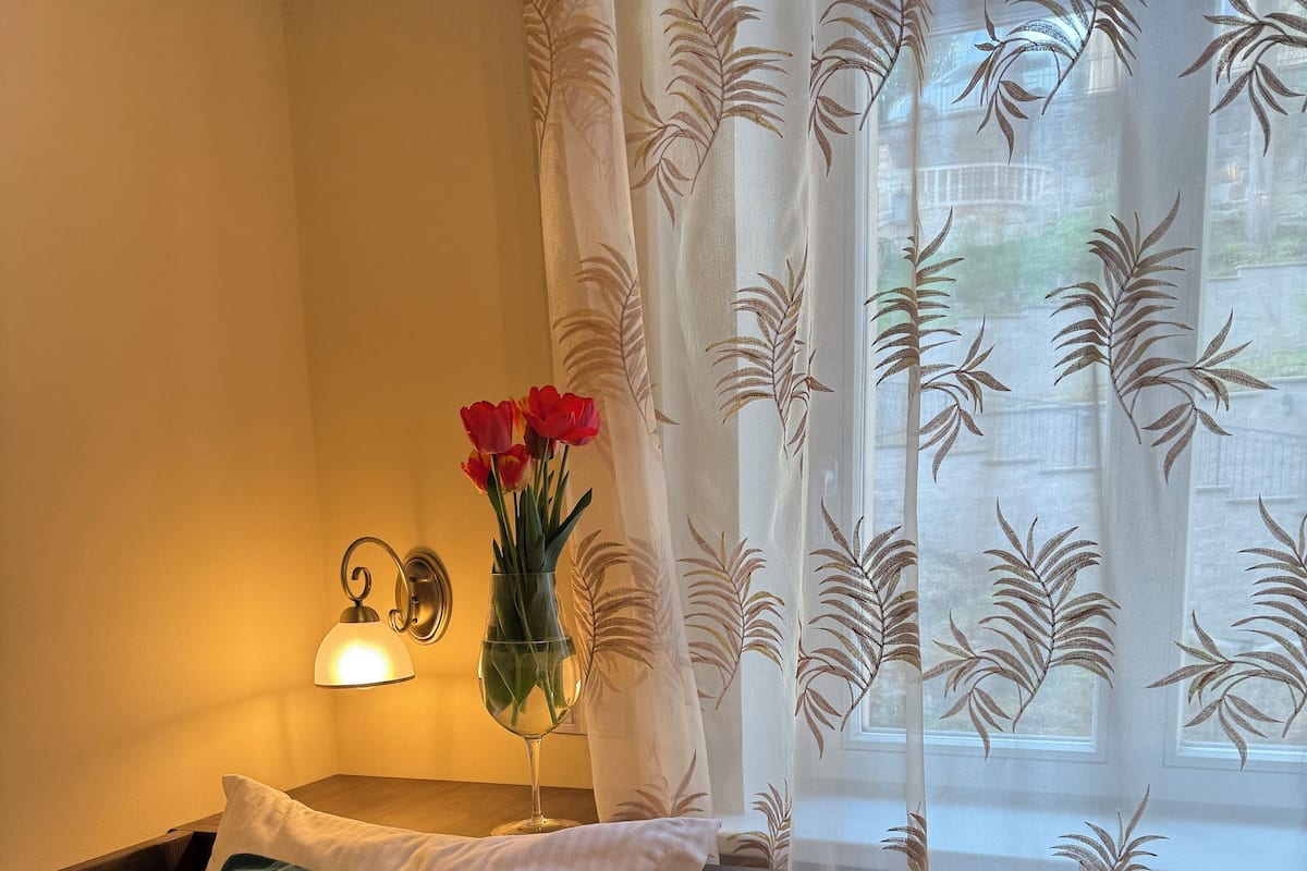 A neatly made bed with a patterned bedspread, a green pillow, and a vase with red flowers on a bedside table.