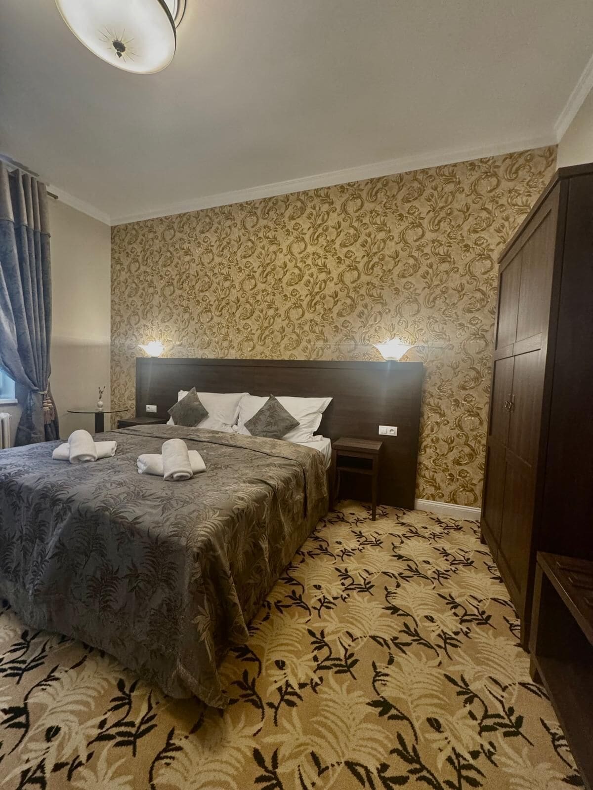 A hotel room with a large bed, a nightstand, a wardrobe, and patterned wallpaper.