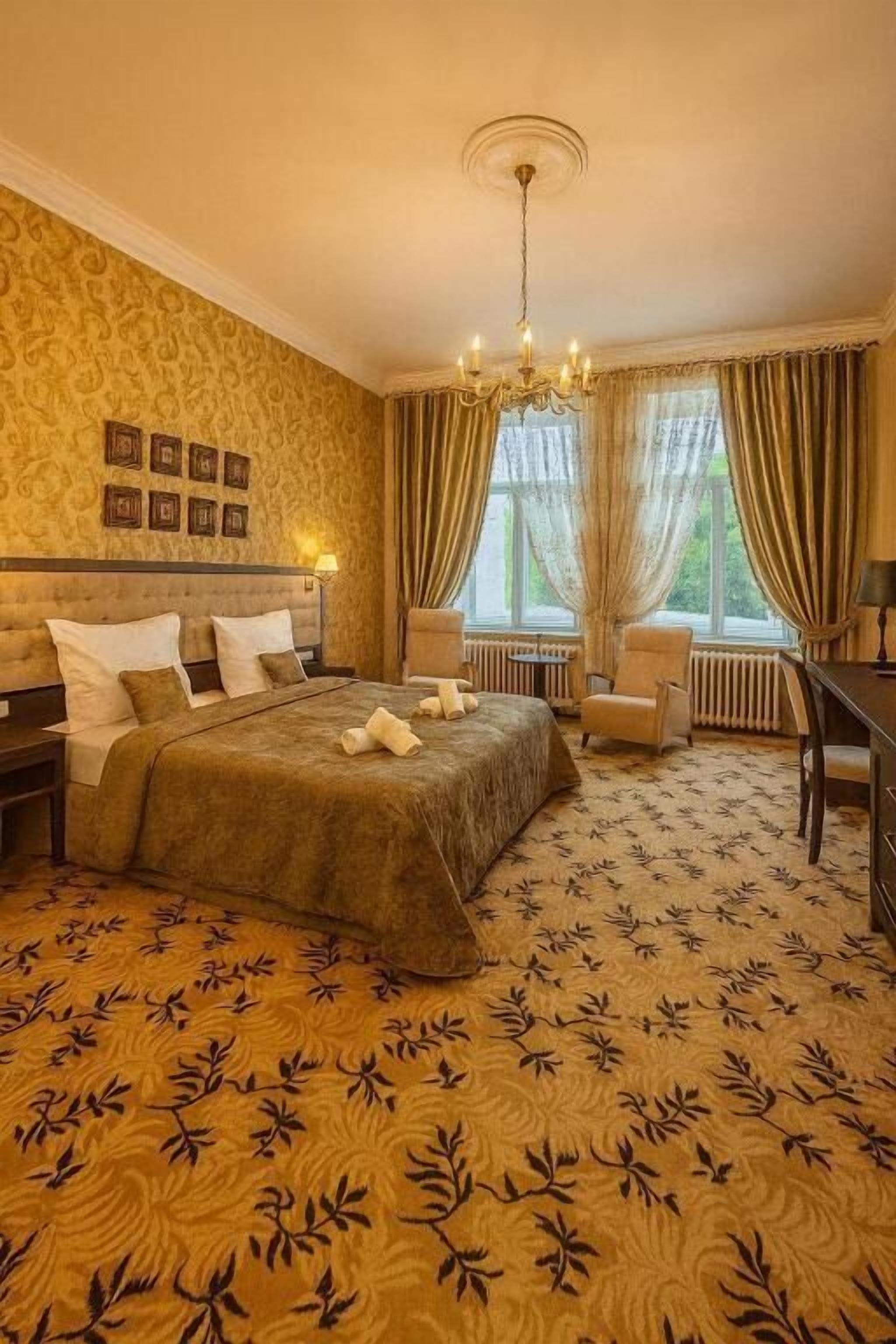 A hotel room with a large bed, two bedside tables, a desk, and a chair. There are curtains on the windows and a chandelier on the ceiling.