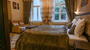 A bedroom with a large bed, a sitting area with a chair and a small table, a radiator, and a window with curtains.