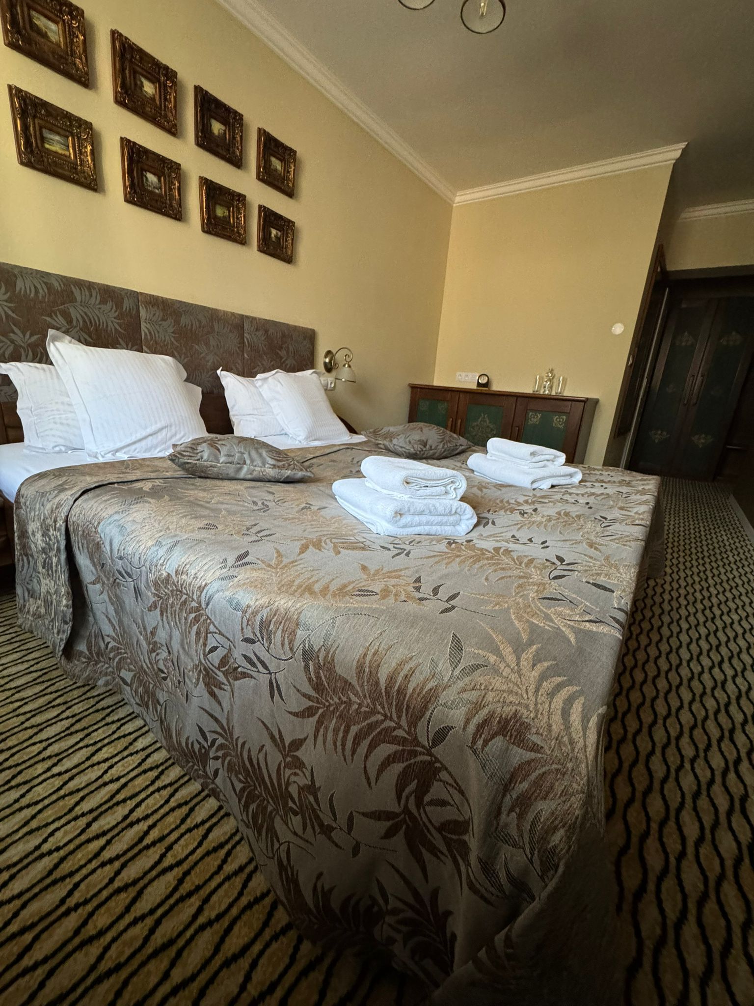 A hotel room with a bed, pillows, a headboard, a wall with framed pictures, a nightstand, and a wardrobe.