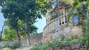Un mur de pierre orné de verdure, une terrasse en bois et un bâtiment avec des colonnes en arrière-plan.