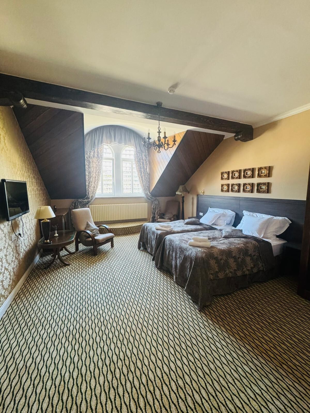 A bedroom with two beds, a desk, a chair, a window with curtains, and a chandelier.