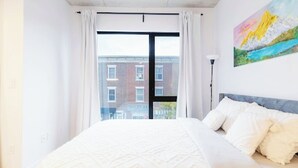 A bedroom with a large bed, white bedding, a painting on the wall, and a view of buildings outside. - Lovely studio for rent (Montréal)