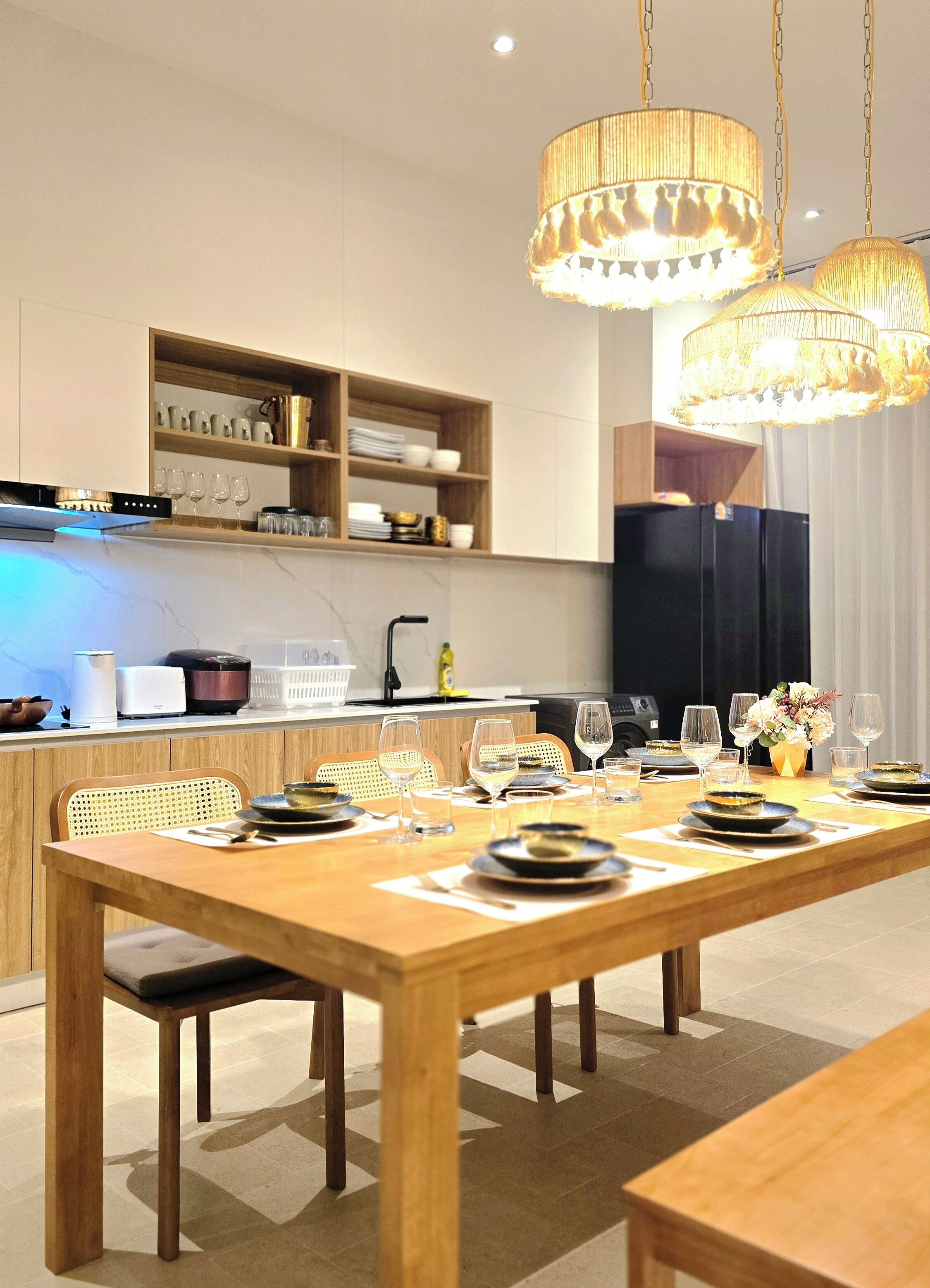 A modern kitchen with wooden cabinets, a dining table set for six, and hanging pendant lights.
