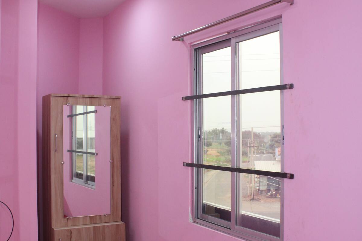 A bedroom with pink walls, a wooden wardrobe, a bed with a red patterned bedspread, and a window with metal bars.