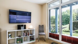 Un salon moderne avec un téléviseur fixé au mur qui affiche une liste de programmes télévisés, une étagère blanche contenant des livres et des objets de décoration, un fauteuil gris et une grande fenêtre donnant sur un paysage verdoyant.