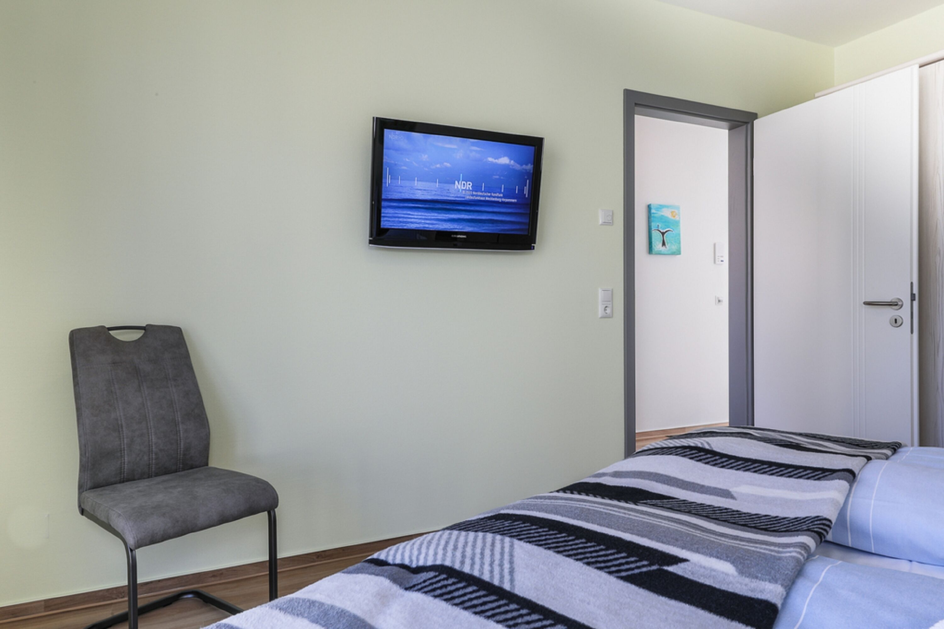 A room with a bed, a chair, a television on the wall, and a door.
