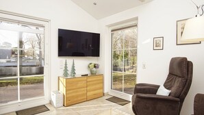 A modern living room with a large flat-screen TV mounted on the wall, a brown leather armchair, a wooden cabinet with decorative items, and a large window offering a view of the outdoors. - Rügen vacation near the beach - Landhaus Wilkens Bungalow Nordlicht (Baabe)