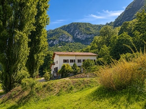 Un bĂątiment Ă deux Ă©tages, dotĂ© dâun toit en pente, entourĂ© dâune vĂ©gĂ©tation luxuriante et avec des montagnes en arriĂšre-plan.