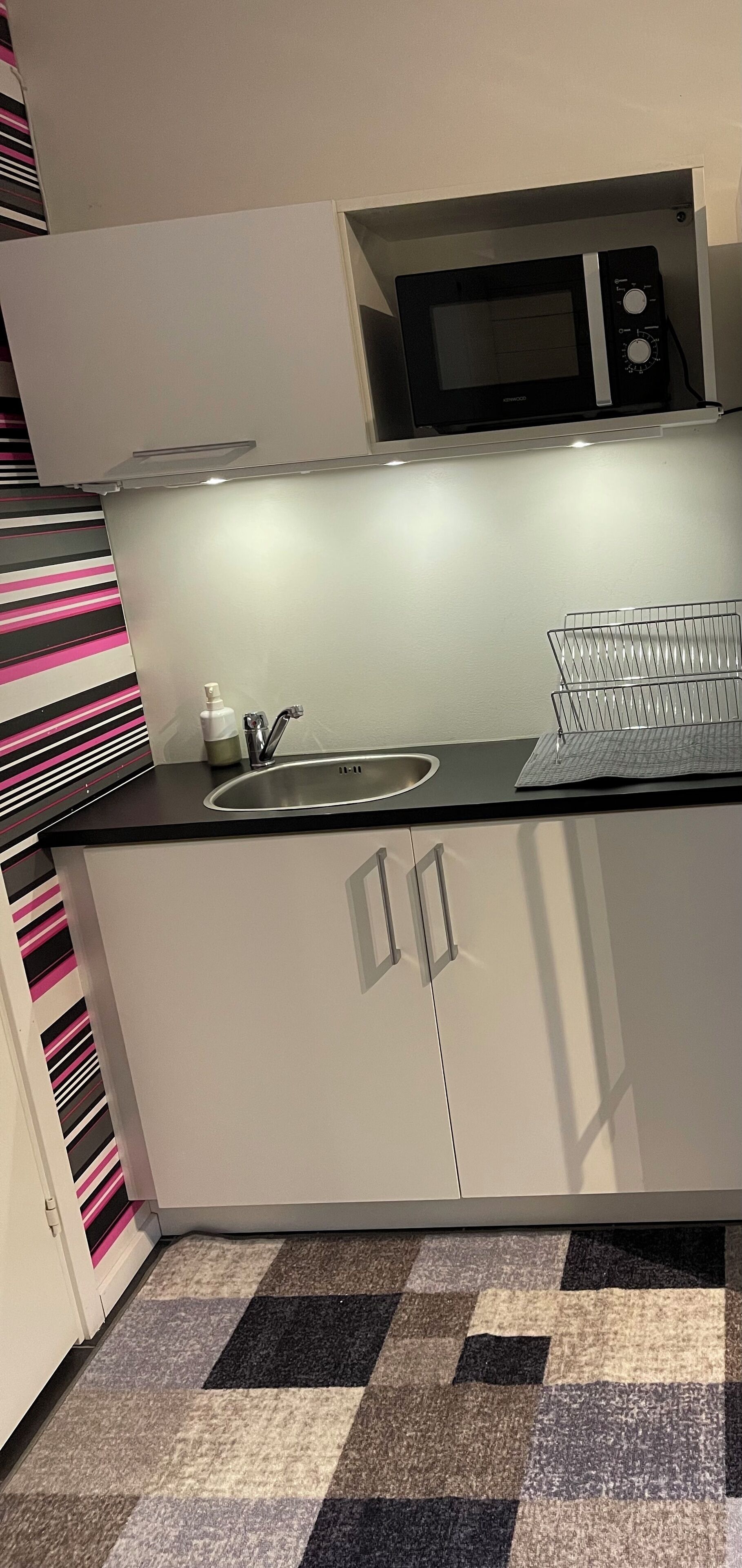 A compact kitchen with a microwave, sink, and a metal drying rack.