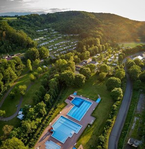 Eine Vogelperspektive zeigt einen Campingplatz mit mehreren Wohnmobilstellplätzen, einem Schwimmbad und bewaldeten Hügeln im Hintergrund.